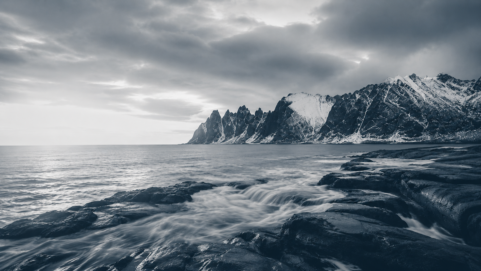 Muted blue-grey seascape with jagged snow-covered mountains across the water